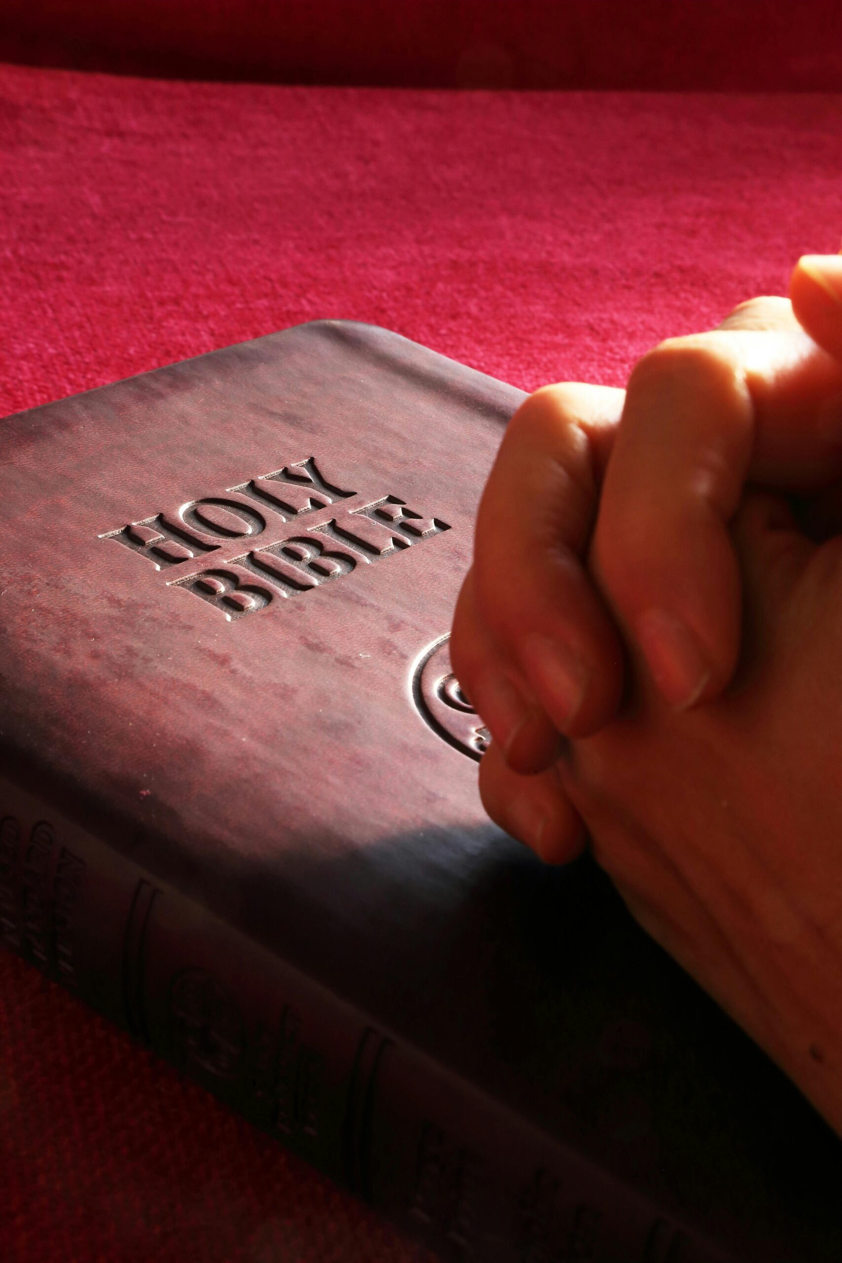 Close-up of clasped hands in prayer resting on a Holy Bible with a dark leather cover embossed with the words 'HOLY BIBLE,' placed against a red background.