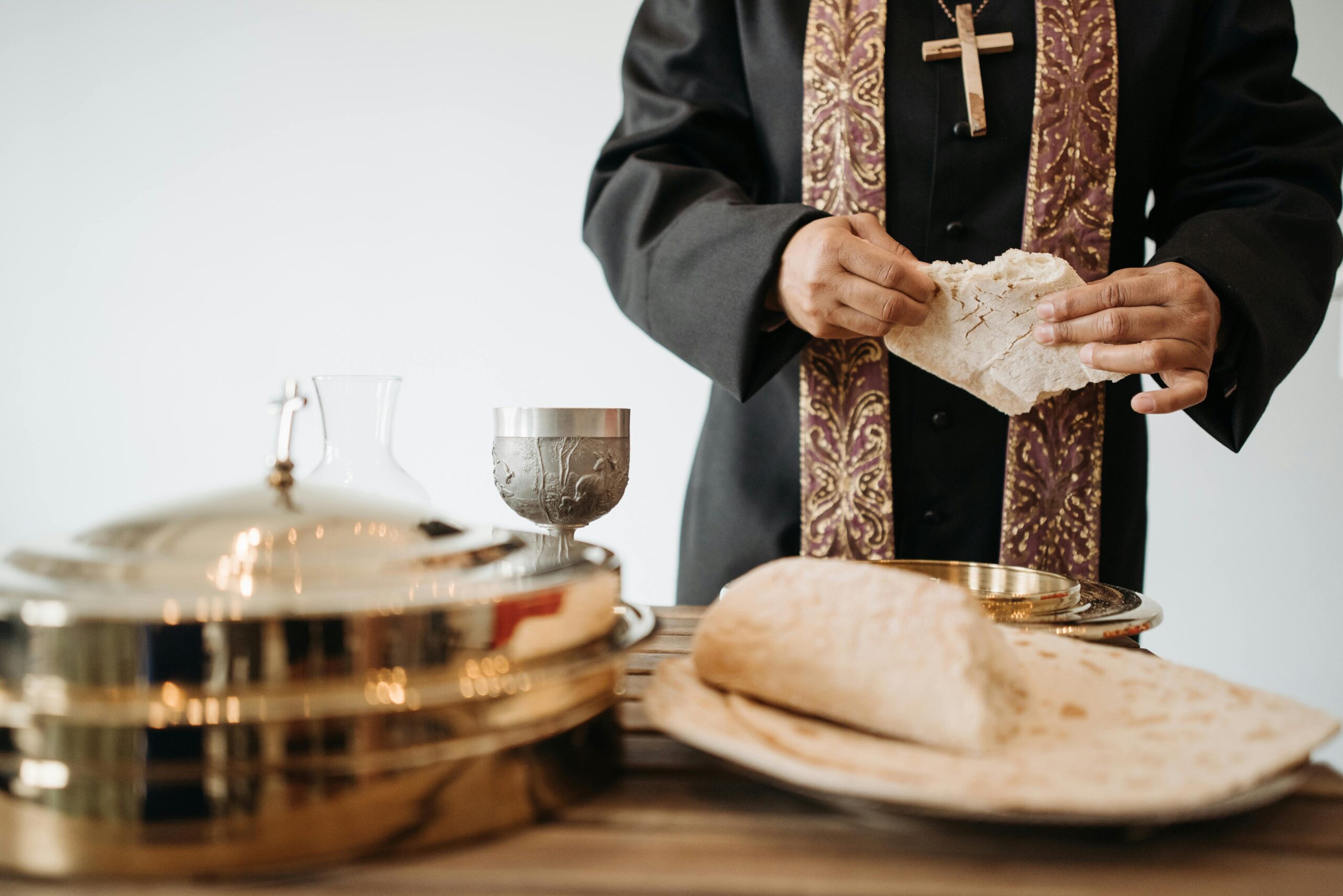 A clergy member in ceremonial attire with a cross necklace breaks a piece of bread during a communion service. The table holds a silver chalice, a glass carafe, and golden communion trays, alongside flatbread, symbolizing the Eucharist. The setting conveys a solemn and sacred atmosphere.