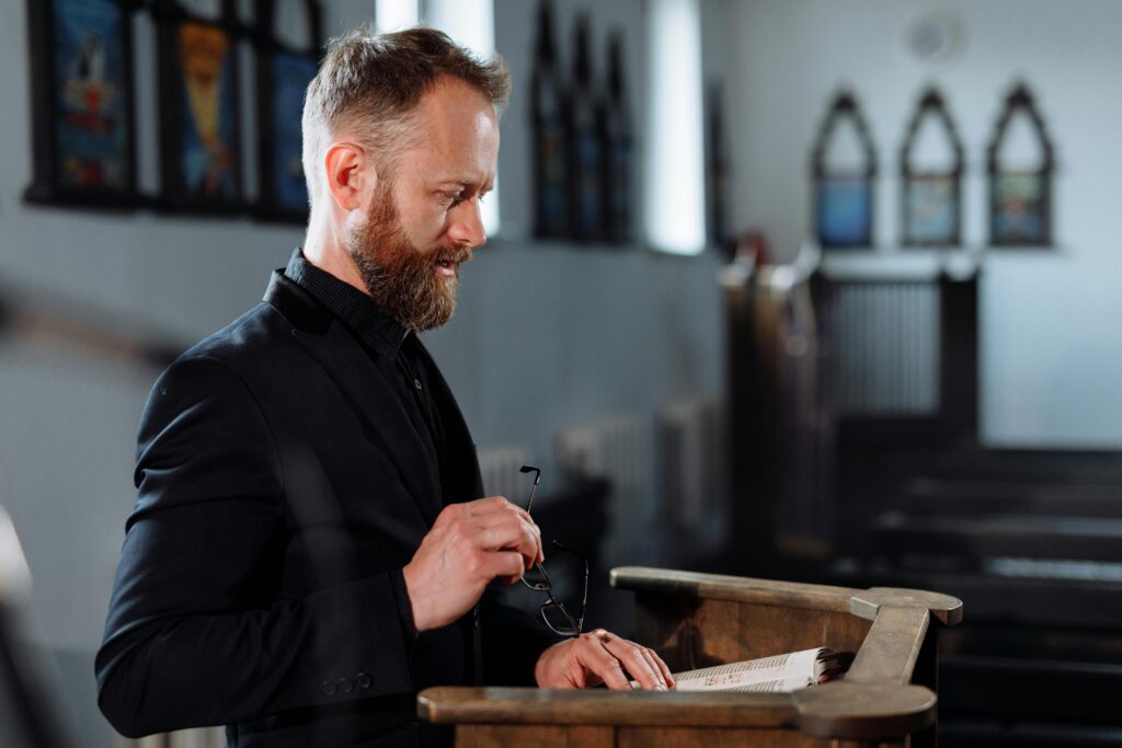 A church leader standing at a wooden pulpit, holding glasses in one hand while referencing an open Bible. The setting includes softly lit stained glass windows and wooden pews, creating a reflective and reverent atmosphere.