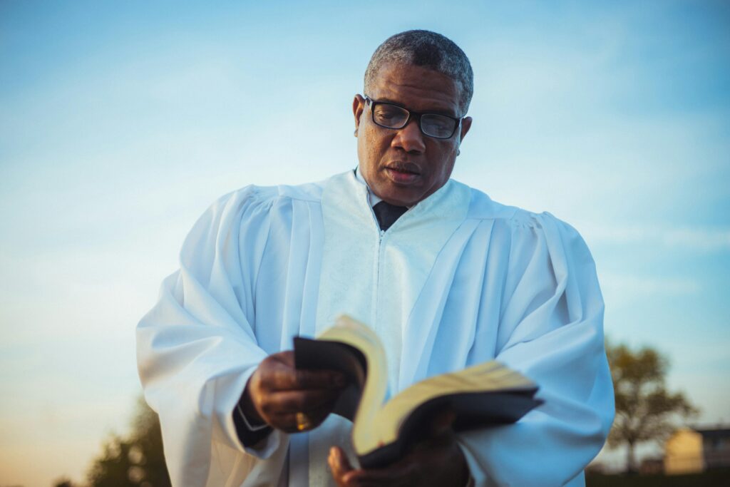 A pastor wearing white liturgical robes reads from an open Bible outdoors under a clear blue sky. The serene background includes trees and soft sunlight, evoking a sense of reflection and spiritual leadership.