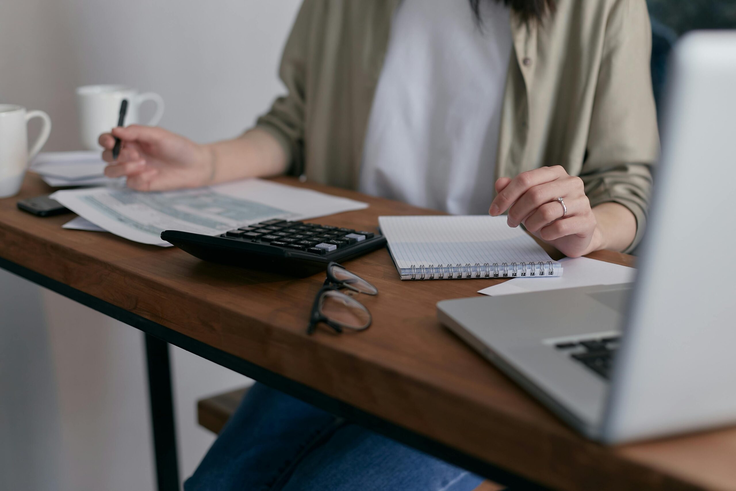 A person seated at a wooden desk, surrounded by a calculator, papers, a notebook, glasses, and a laptop. The scene conveys focus and productivity, possibly related to financial planning or budgeting, with a calm and organized atmosphere.