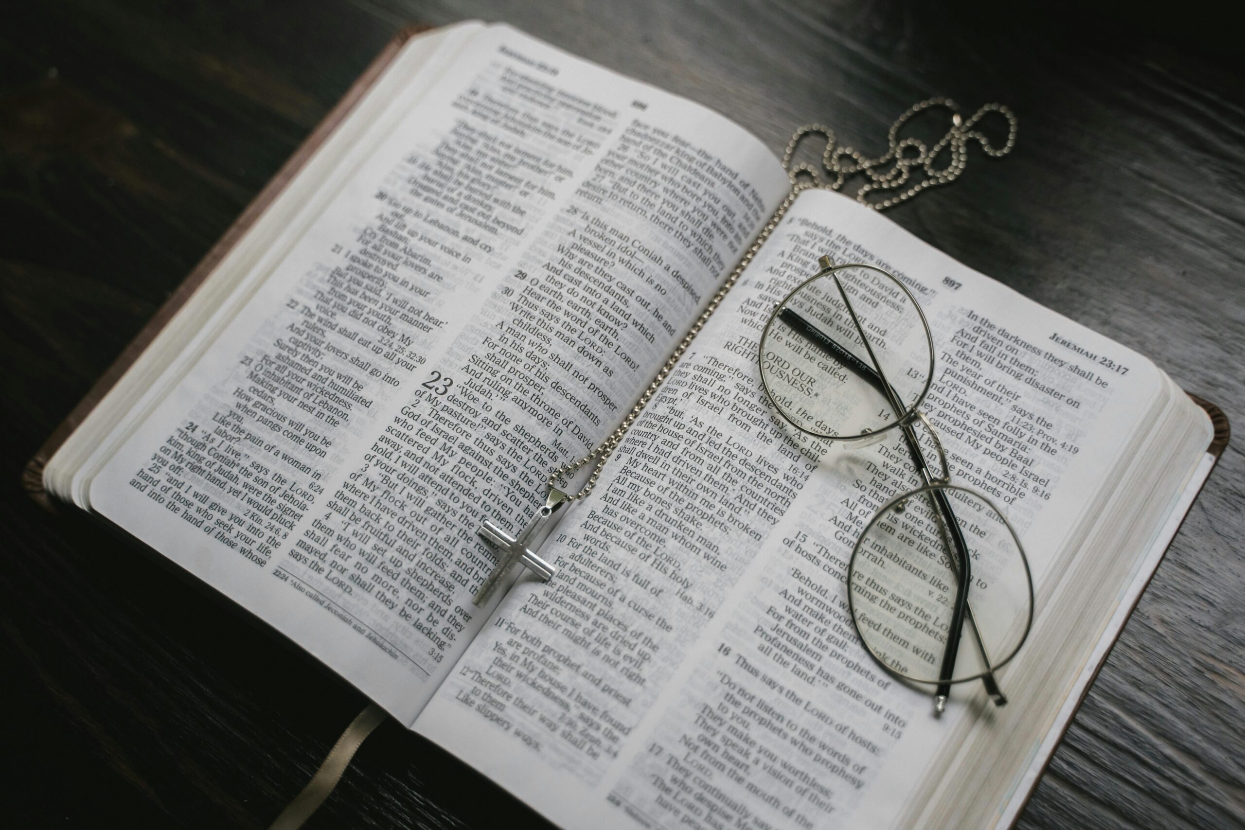 An open Bible on a dark wooden table with a pair of round eyeglasses and a silver cross necklace resting on the pages. The Bible is open to passages in the Old Testament, with a ribbon bookmark peeking out below, evoking a sense of contemplation and faith.