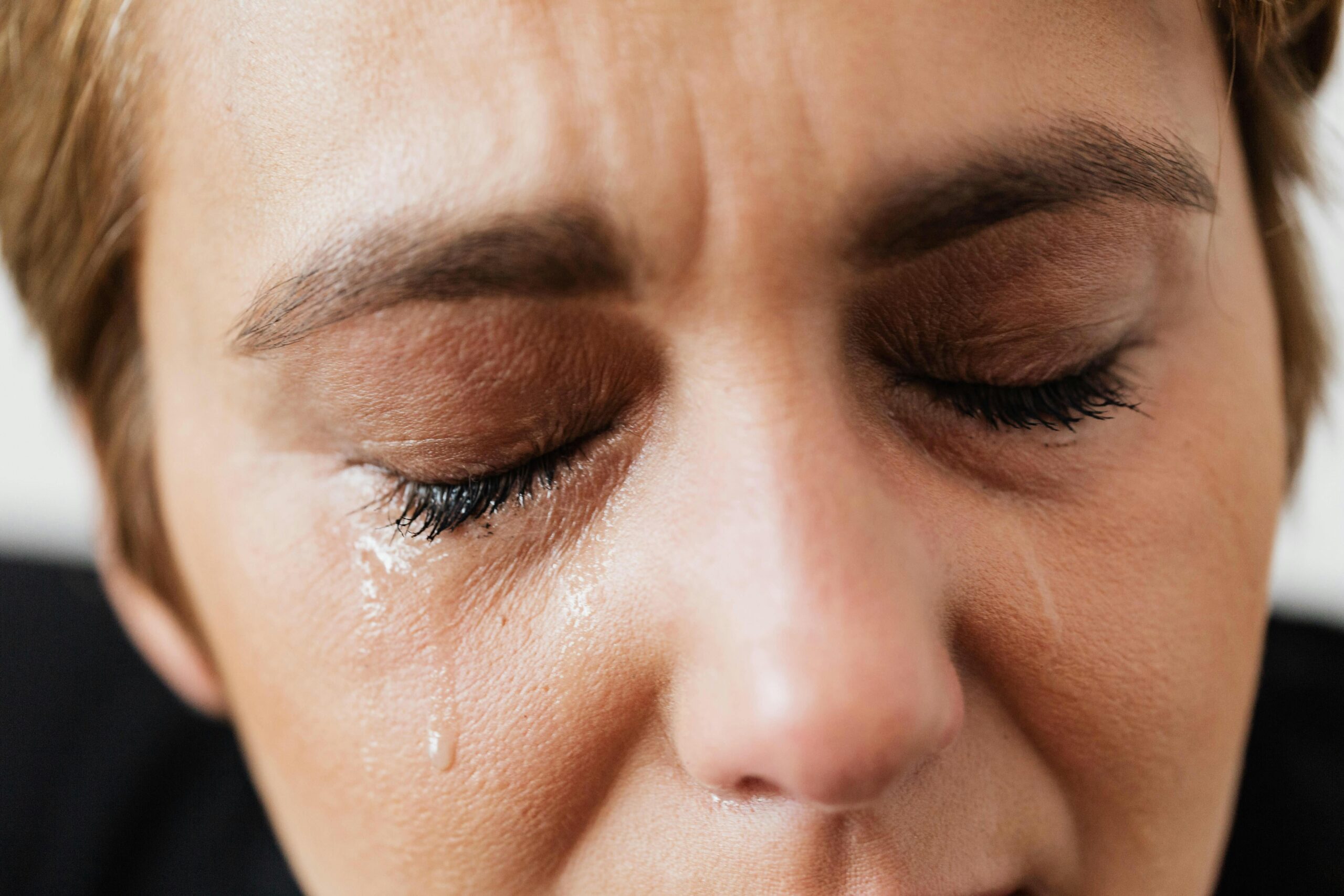 Close-up of a woman with closed eyes and tears streaming down her face, conveying deep emotion or grief. The image highlights the raw and intimate expression of sorrow, with fine details of the eyelashes and skin texture.