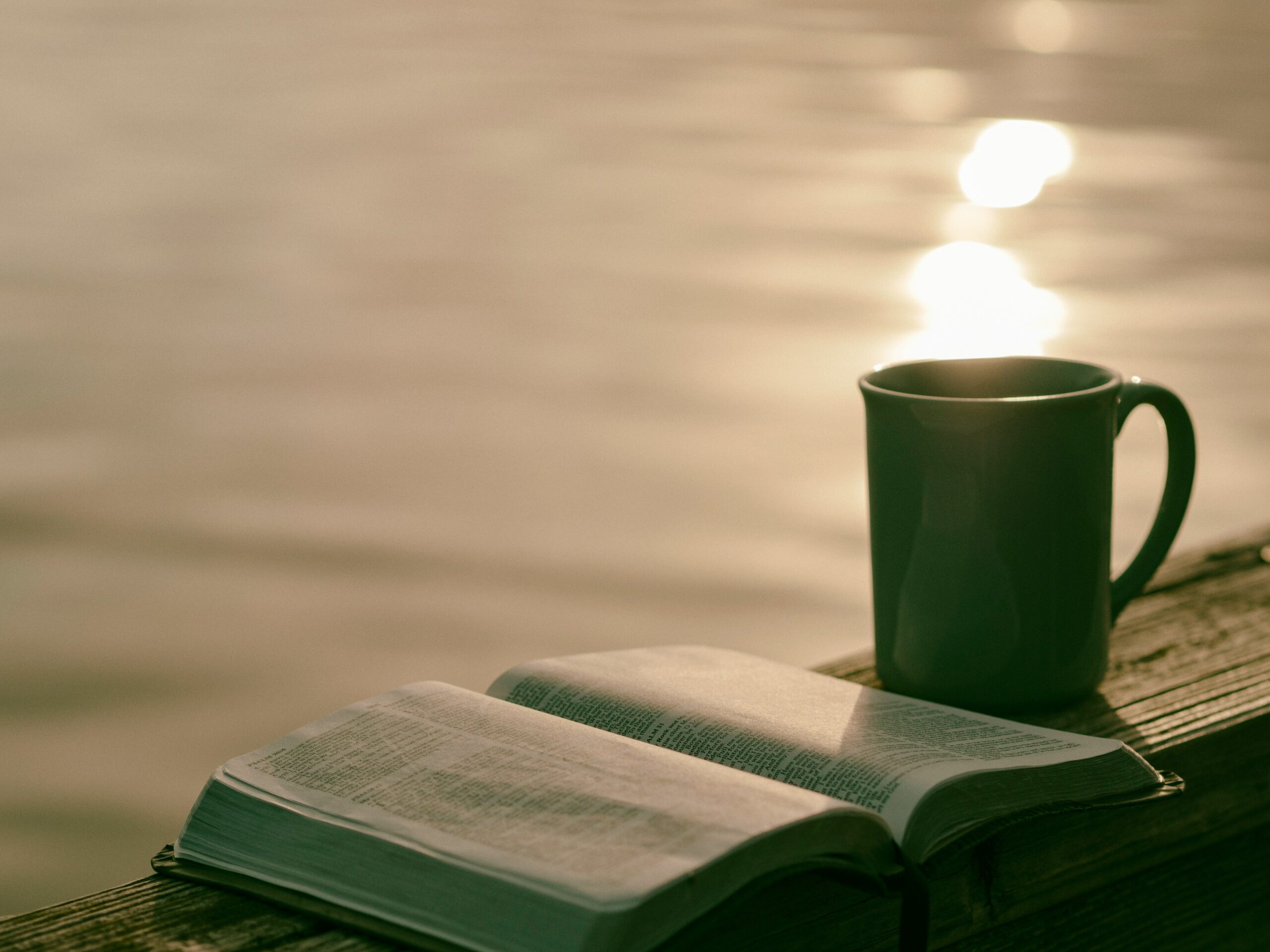 An open Bible and a mug resting on a wooden surface by a calm body of water during sunrise or sunset. The golden light reflects off the water, creating a peaceful and reflective atmosphere, ideal for morning devotion or quiet meditation.