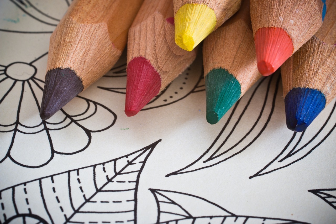 Close-up of colorful wooden pencils, including red, yellow, green, and blue, resting on a black-and-white coloring page with intricate floral and leaf designs. The image conveys creativity and relaxation, suitable for adult coloring activities.