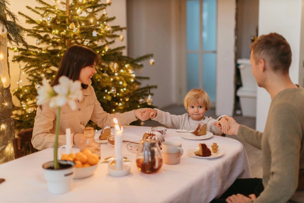 A family of three holds hands around a dining table in prayer, with a decorated Christmas tree glowing softly in the background. The table is set with candles, slices of cake, and festive decor, creating a warm and serene holiday atmosphere.