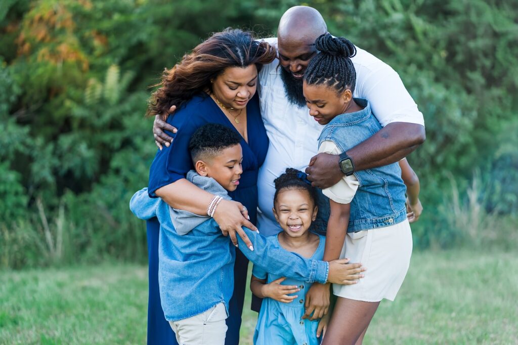 A family of five standing outdoors embraces each other in a group hug, smiling and sharing a joyful moment. The background features lush green trees and grass, enhancing the warmth and connection in the scene.