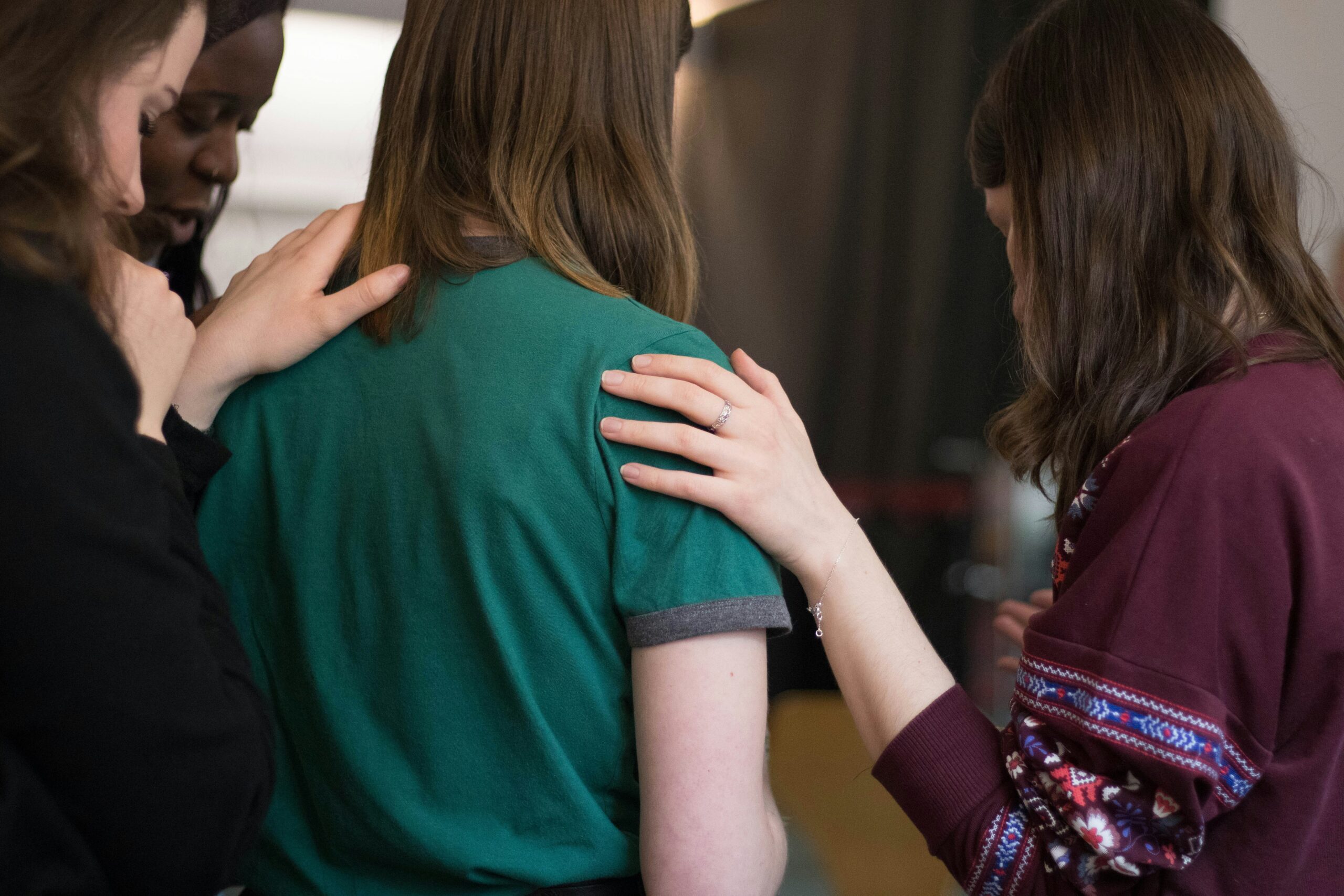 A group of women stands together in a supportive and prayerful moment, with hands placed gently on one person's shoulders. The image captures a sense of encouragement, community, and compassion.