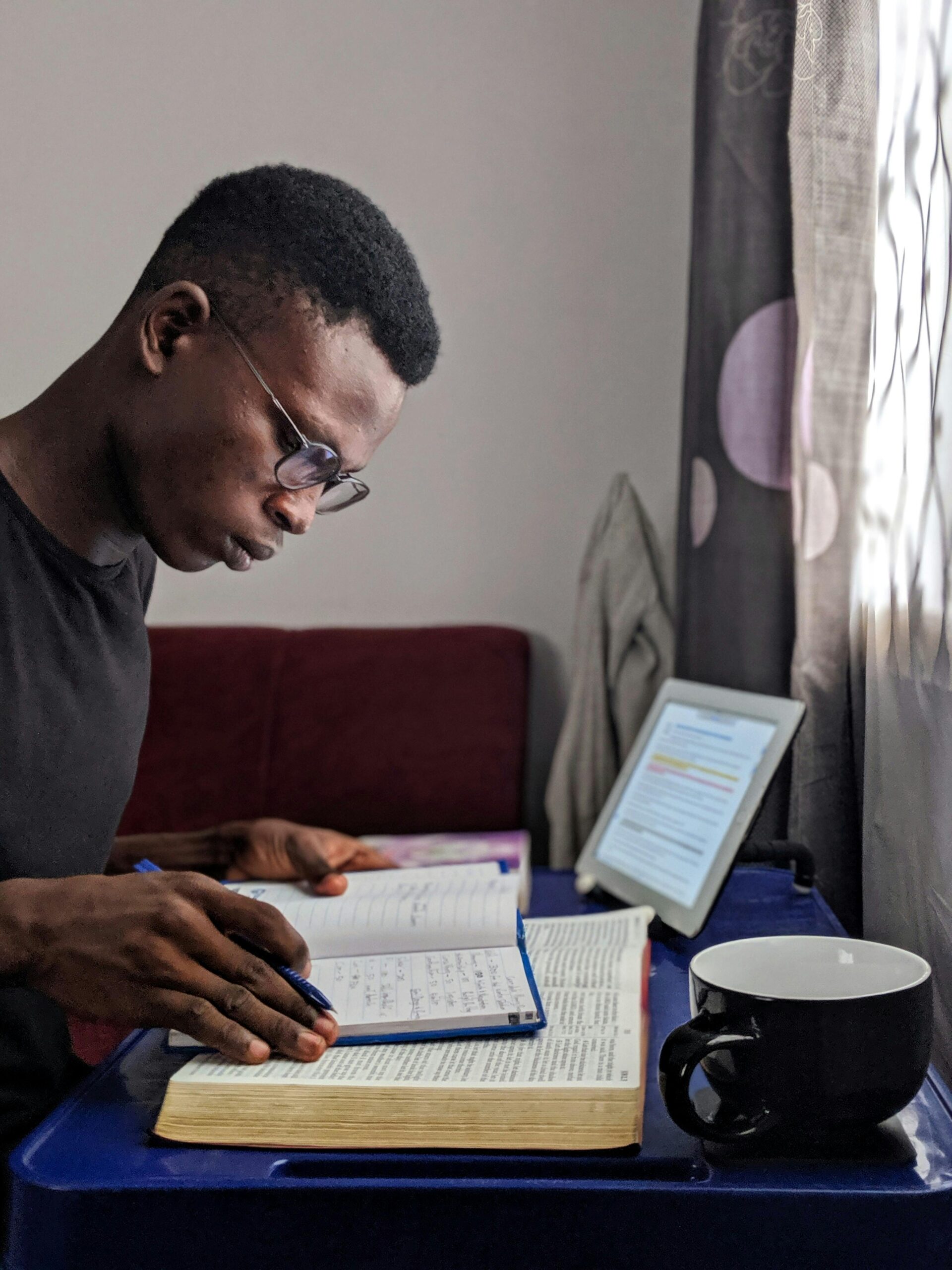 A man wearing glasses is deeply focused on Bible study, seated at a desk with an open Bible, a notebook, and a pen. A tablet displaying highlighted text is propped up nearby, and a black mug sits on the desk, creating an atmosphere of concentration and devotion.