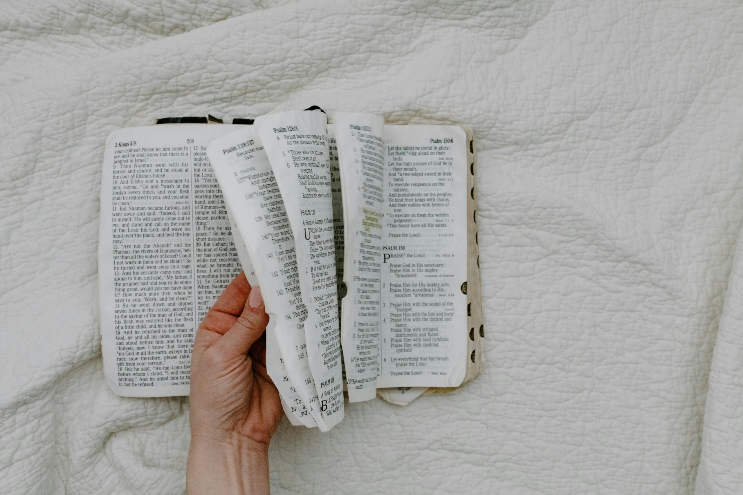 A hand holding the pages of an open Bible, revealing passages from 2 Kings and Psalms. The Bible is placed on a soft white quilted background, creating a serene and reflective atmosphere ideal for devotion or study.