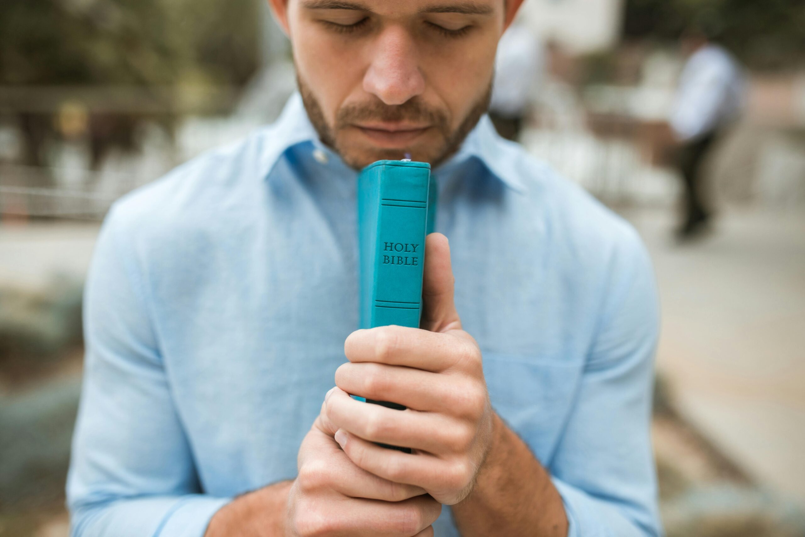 A man in a light blue shirt holds a turquoise Holy Bible close to his face with his eyes closed, appearing to pray or reflect. The blurred outdoor background suggests a serene and contemplative setting, emphasizing faith and personal devotion.