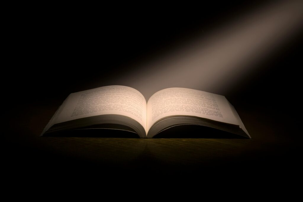 An open book illuminated by a single beam of light in a dark room, emphasizing its pages and text. The dramatic lighting creates a sense of mystery and focus on the content of the book.