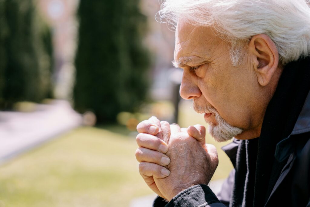 An elderly man with white hair sits outdoors in a park, clasping his hands together in prayer. His expression is reflective and focused, with greenery and blurred trees in the background.