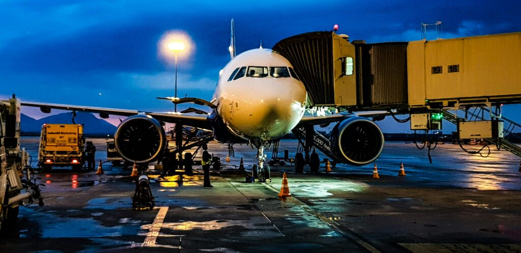 A parked airplane connected to a boarding bridge on a wet tarmac during twilight. Ground crew and vehicles are visible around the aircraft, with soft reflections of lights on the wet ground and a deep blue sky in the background. The scene conveys a sense of travel preparation at an airport.