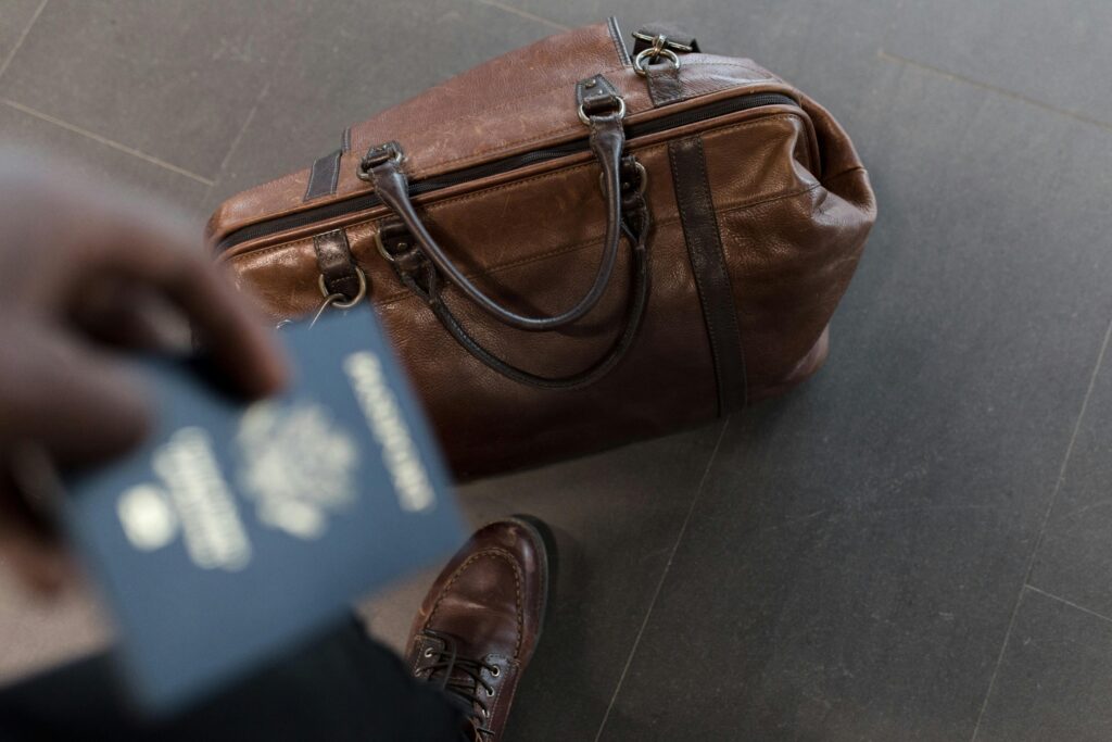 A close-up of a brown leather travel bag on a tiled floor, with a person's hand holding a blurred passport in the foreground. The image suggests preparation for travel, with a polished leather shoe partially visible at the bottom of the frame.